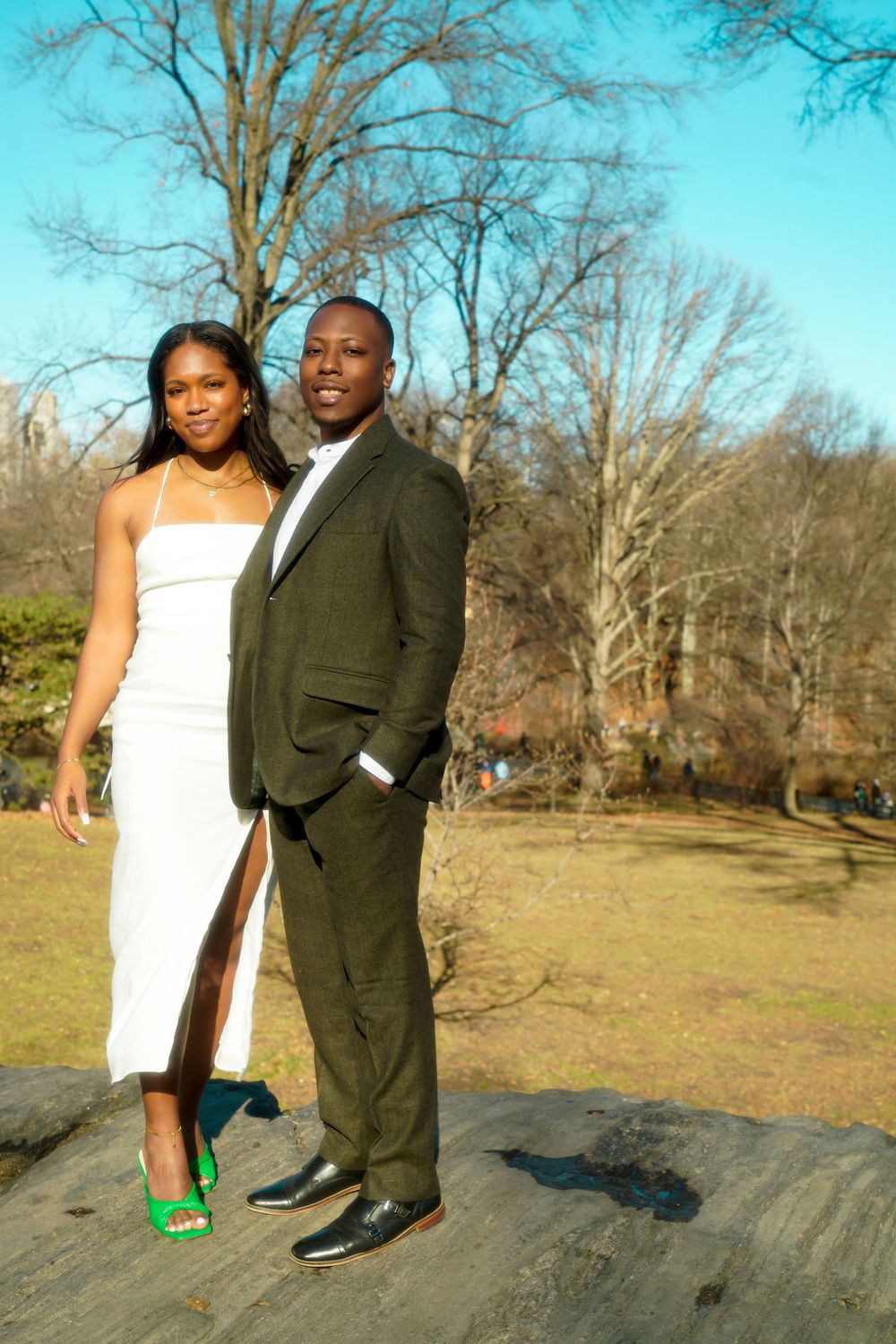 A well-dressed couple poses outdoors in formal attire, with her in a white dress and him in a dark suit. central park