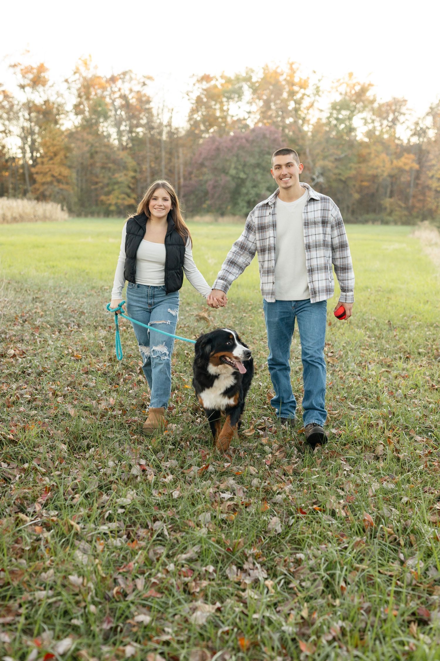 A couple and their black and white dog walk together through a grassy autumn field at sunset.