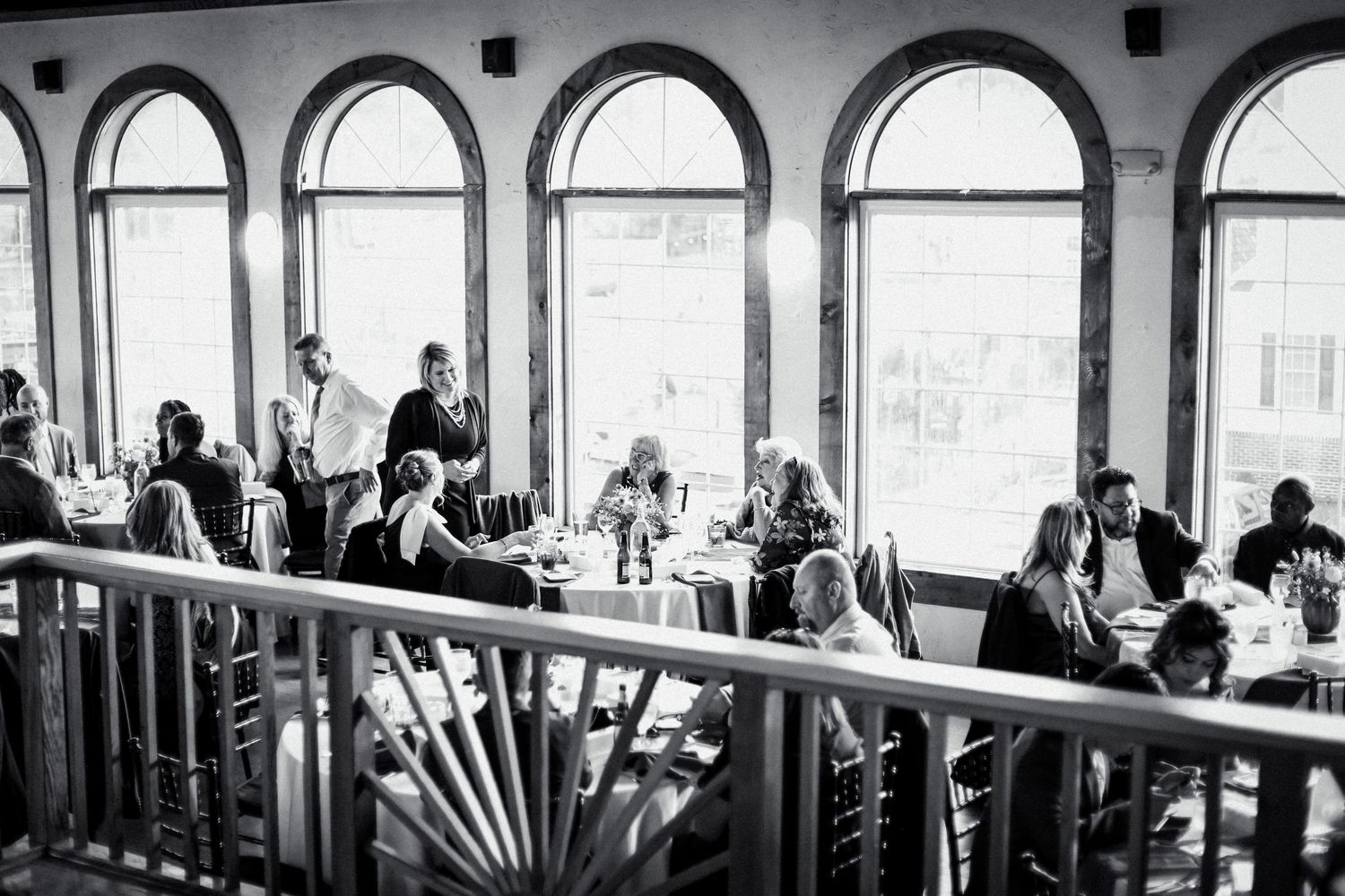 Wedding guests are silhouetted in dramatic black and white while mingling in an elegant reception venue with arched windows.