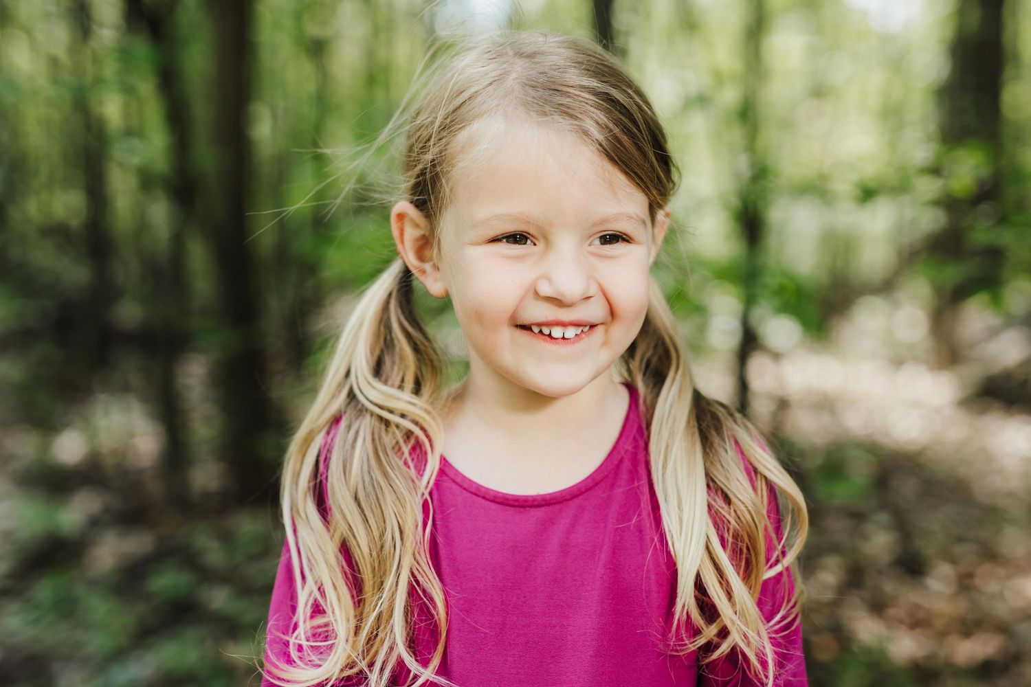 A smiling child with blonde hair wearing a bright pink shirt stands in a sunlit forest setting.