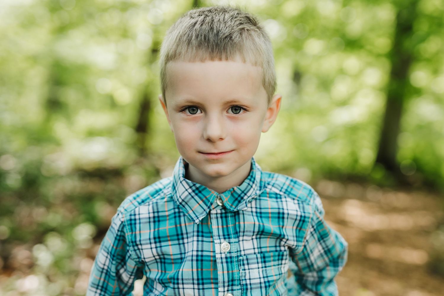 A child wearing a teal and white checkered shirt poses outdoors in a sunlit wooded setting.