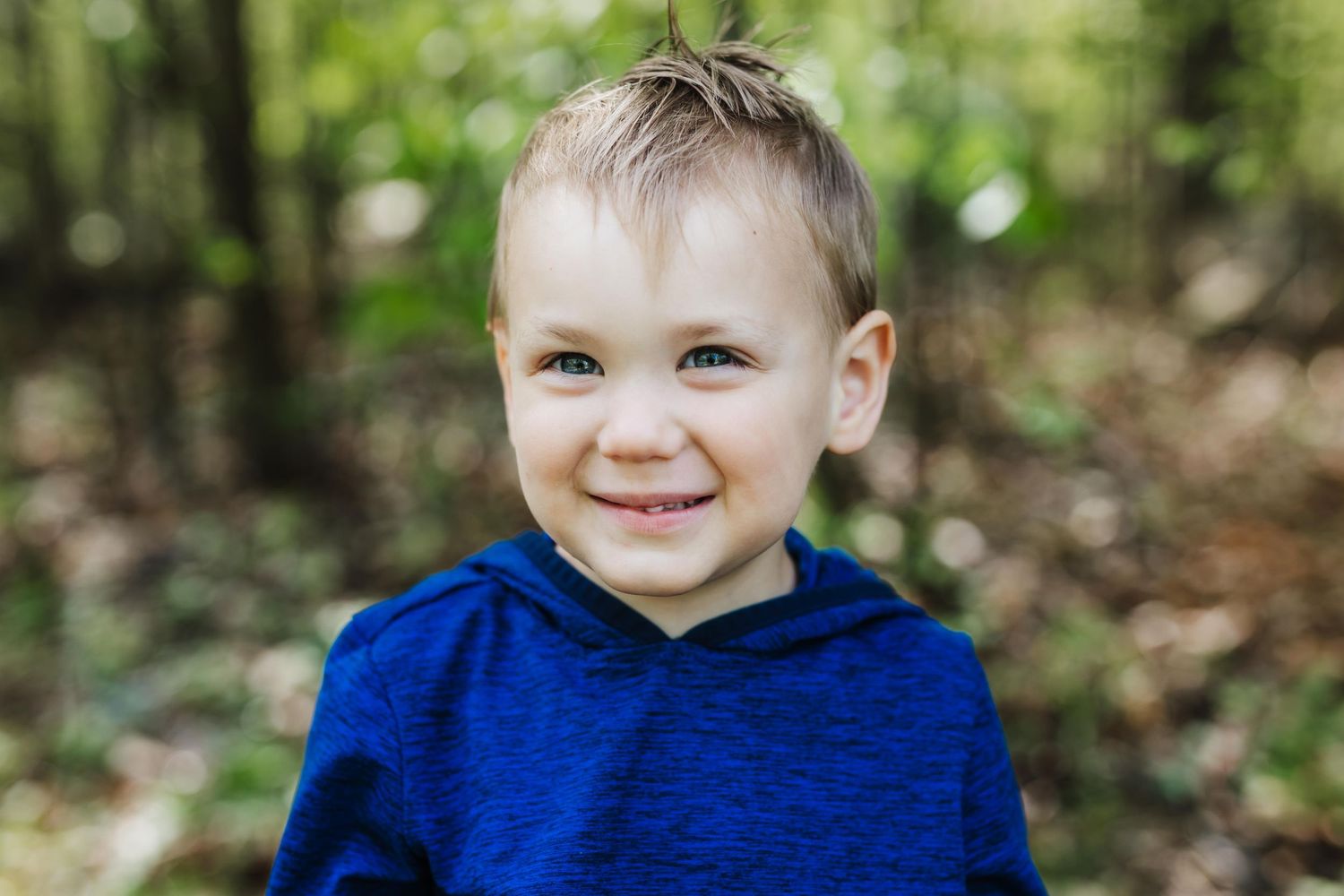 A young child in a bright blue sweater smiling while sitting outdoors against a blurred natural background.