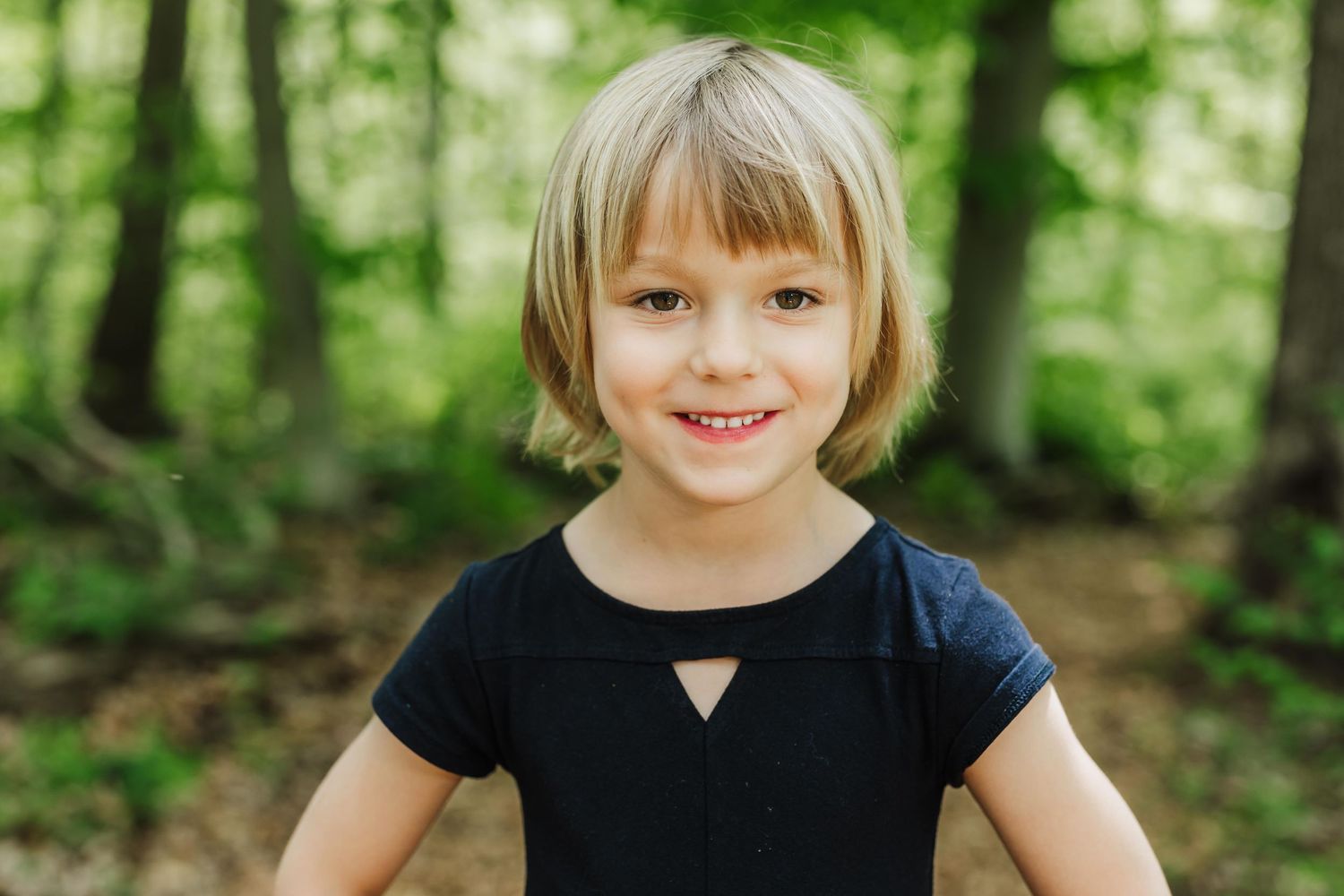 Child wearing a black shirt smiles warmly while standing in a lush green forest setting.