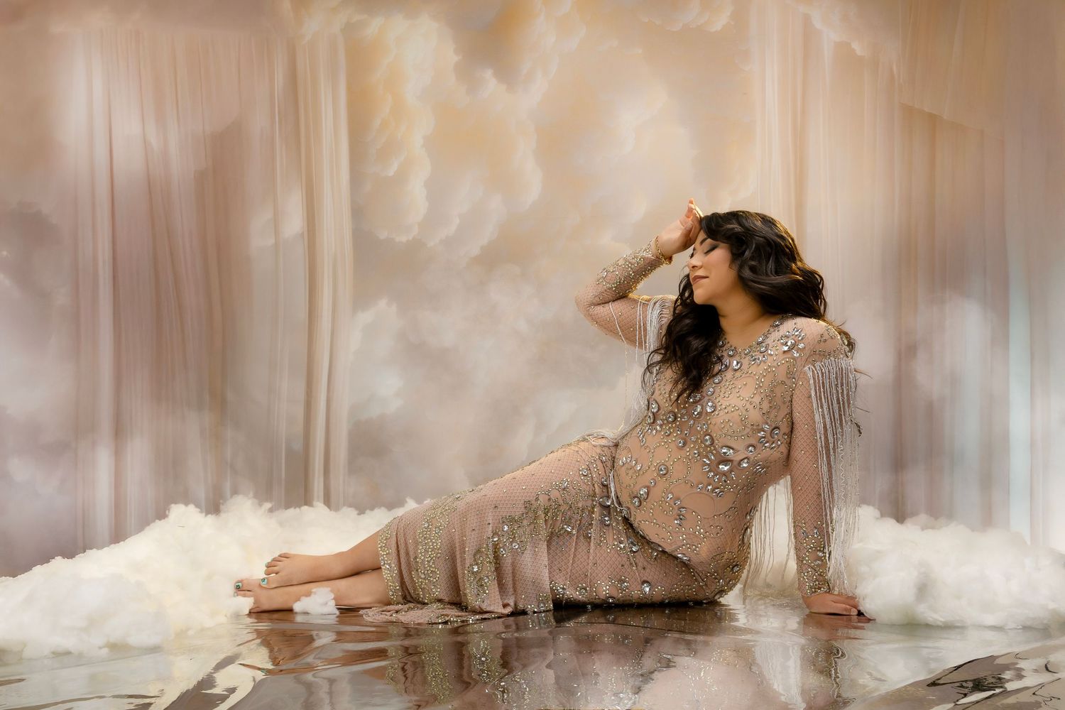 A person in a sparkly dress poses gracefully on a reflective surface surrounded by dreamy smoke effects and white clouds.