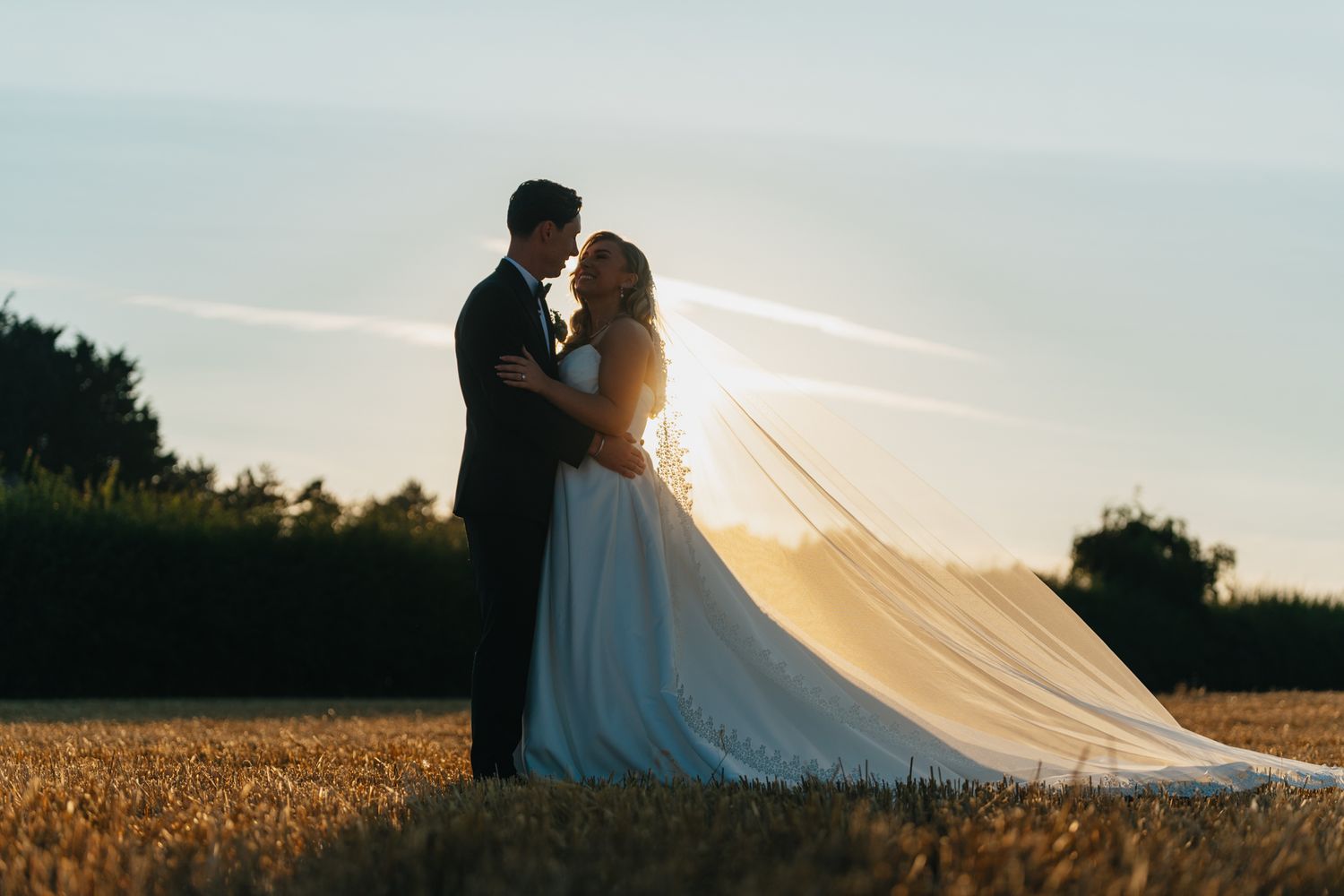 Wedding couple silhouetted against sunset in field with flowing veil catching wind.