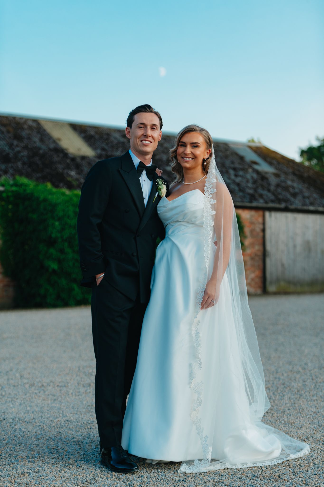 Wedding couple poses against stone wall under bright blue sky during outdoor photo session.