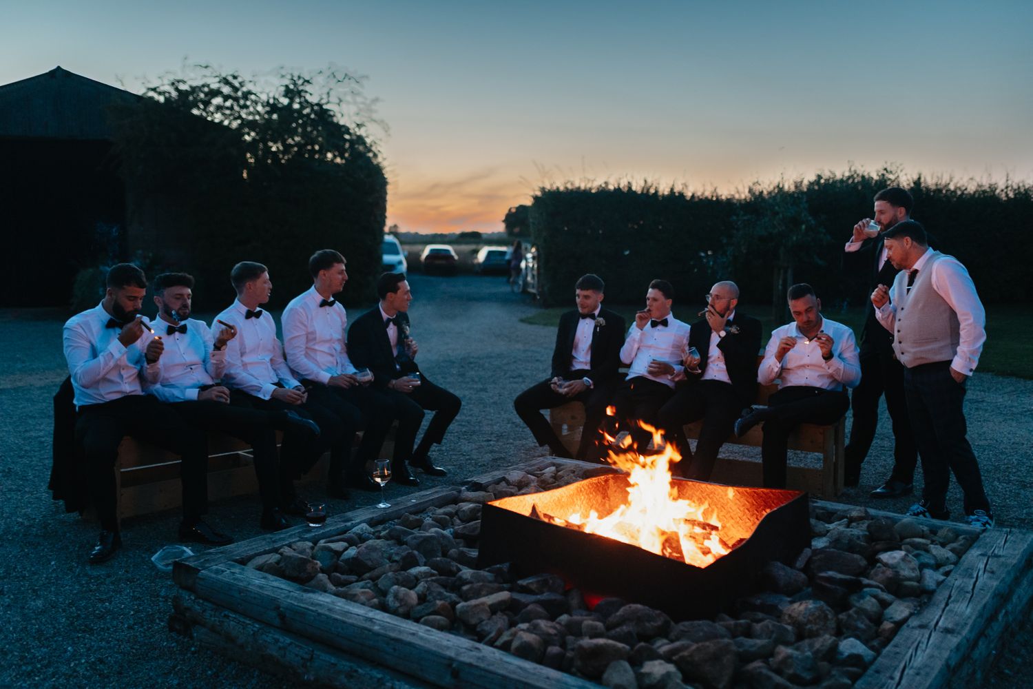 A group gathers around a large square fire pit on a beach at dusk.