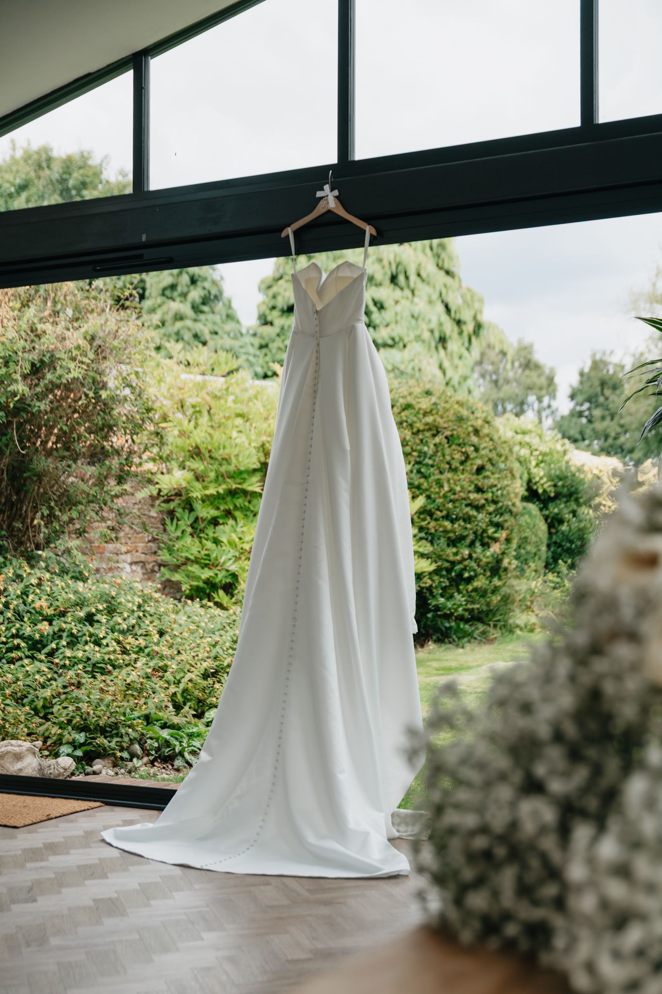 A white wedding dress hangs elegantly against lush green foliage in an outdoor setting.