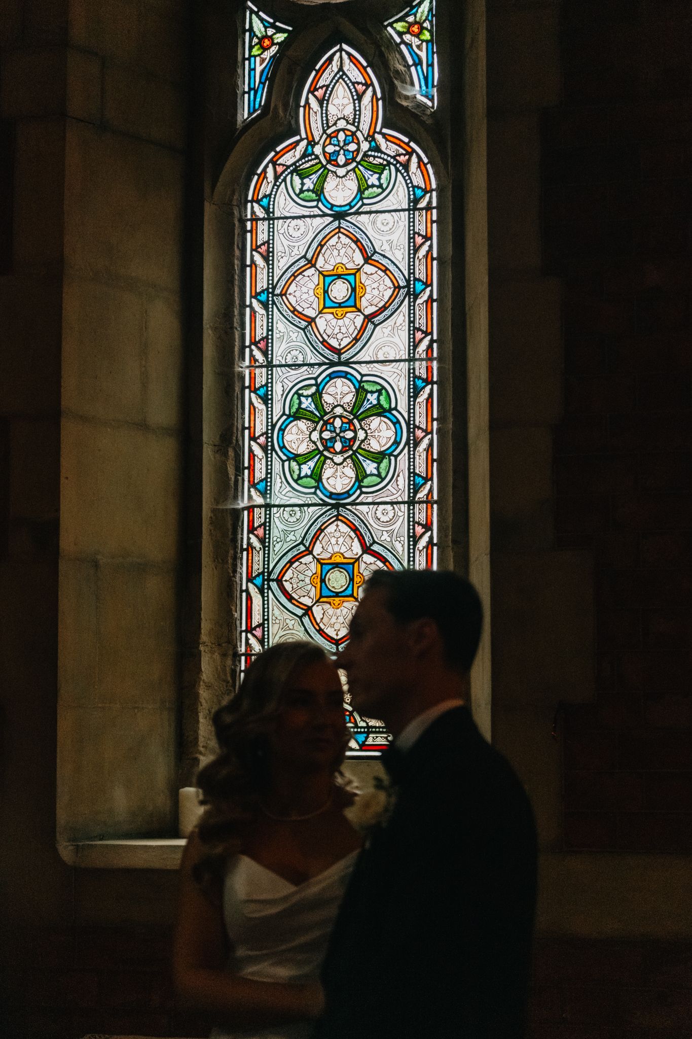 Dramatic silhouettes against ornate stained glass church windows during a wedding ceremony.
