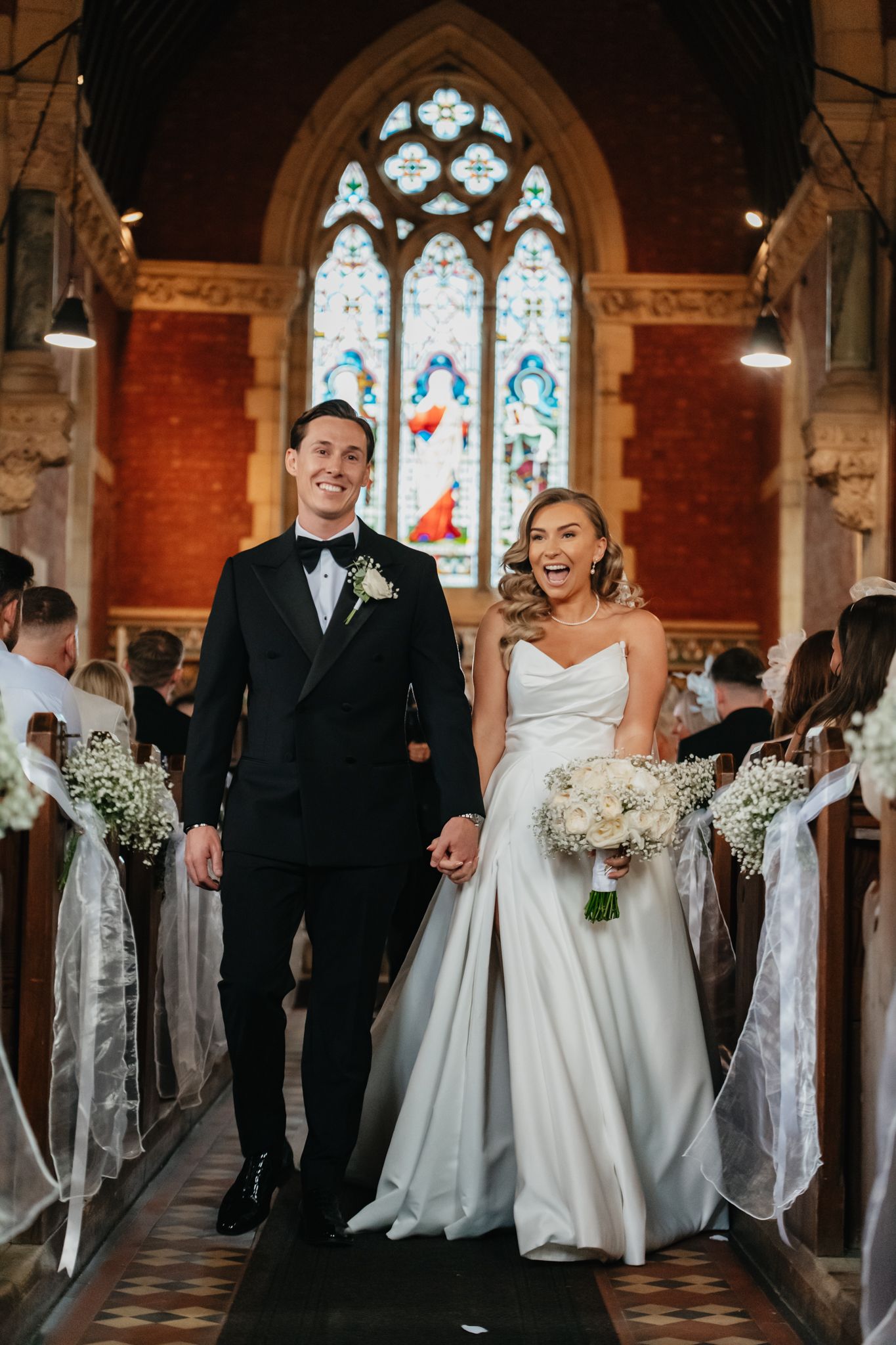 Wedding party walking and laughing in church aisle with stained glass windows and brick walls.