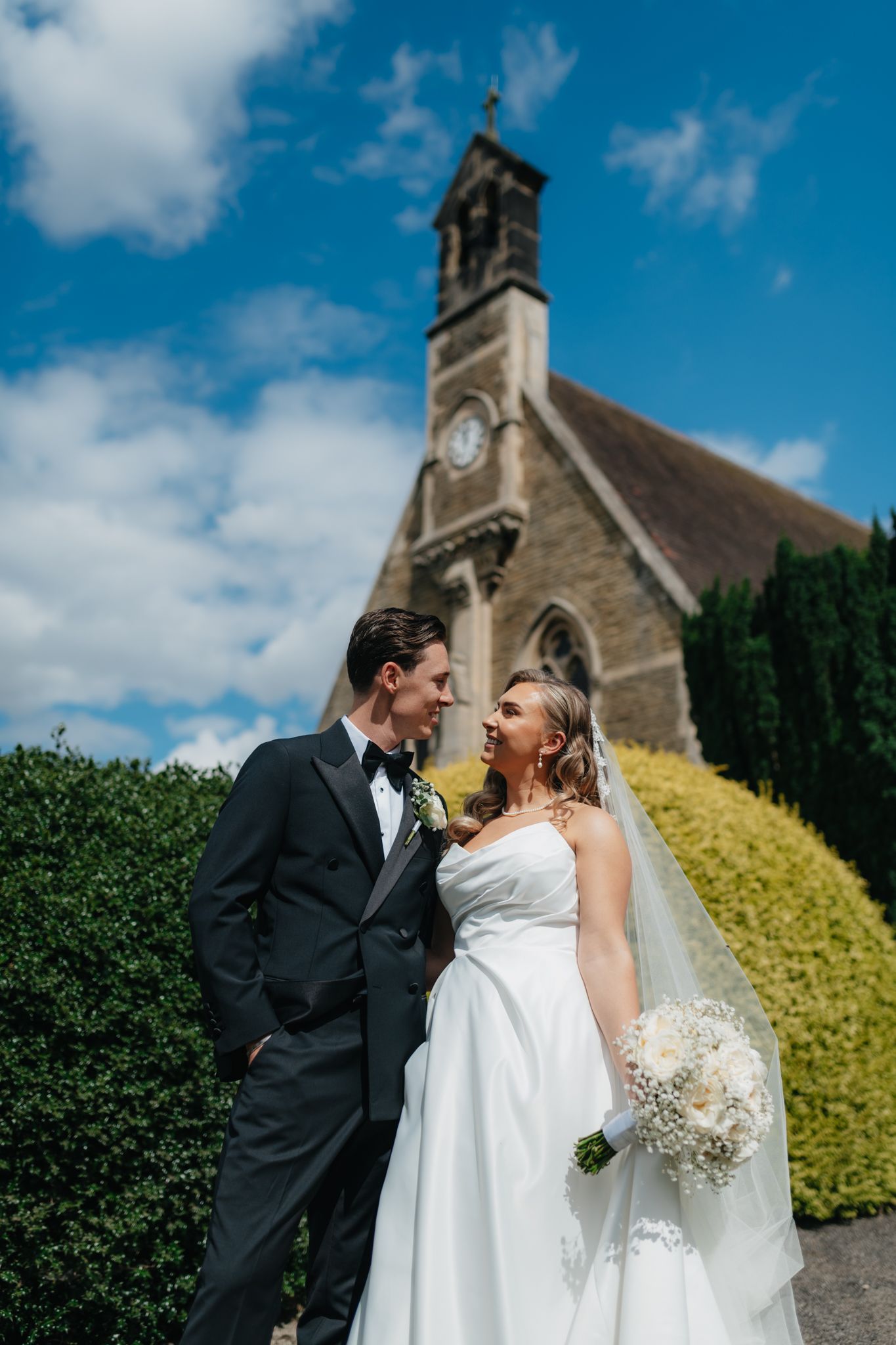 Newlyweds share intimate moment outside church with blue sky and greenery in background.