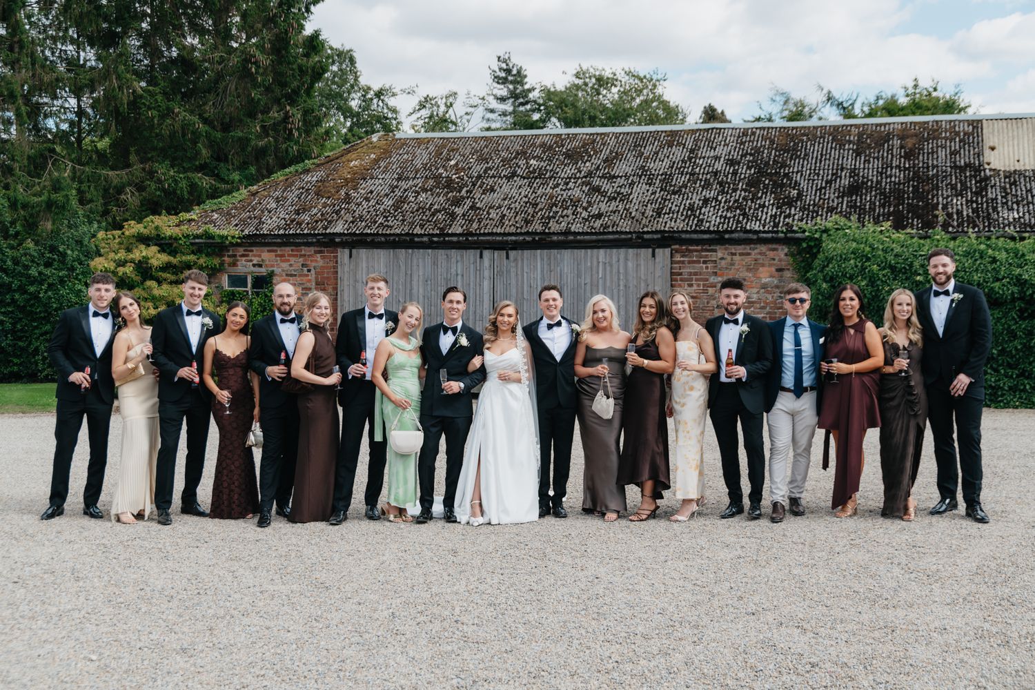 Large wedding party posing outdoors on gravel path with classic building in background.