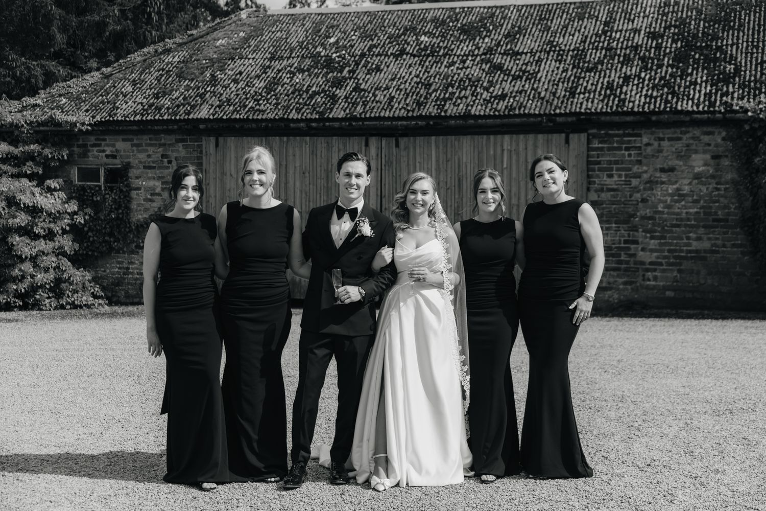 Wedding party in black and white attire lined up against rustic brick wall exterior.