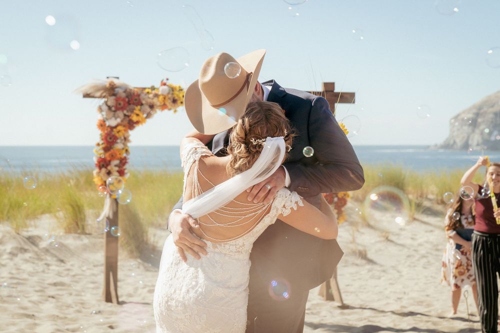 Couple kissing under floral arch with bubbles at Oregon Coast beach wedding – Pacific Northwest wedding photography.