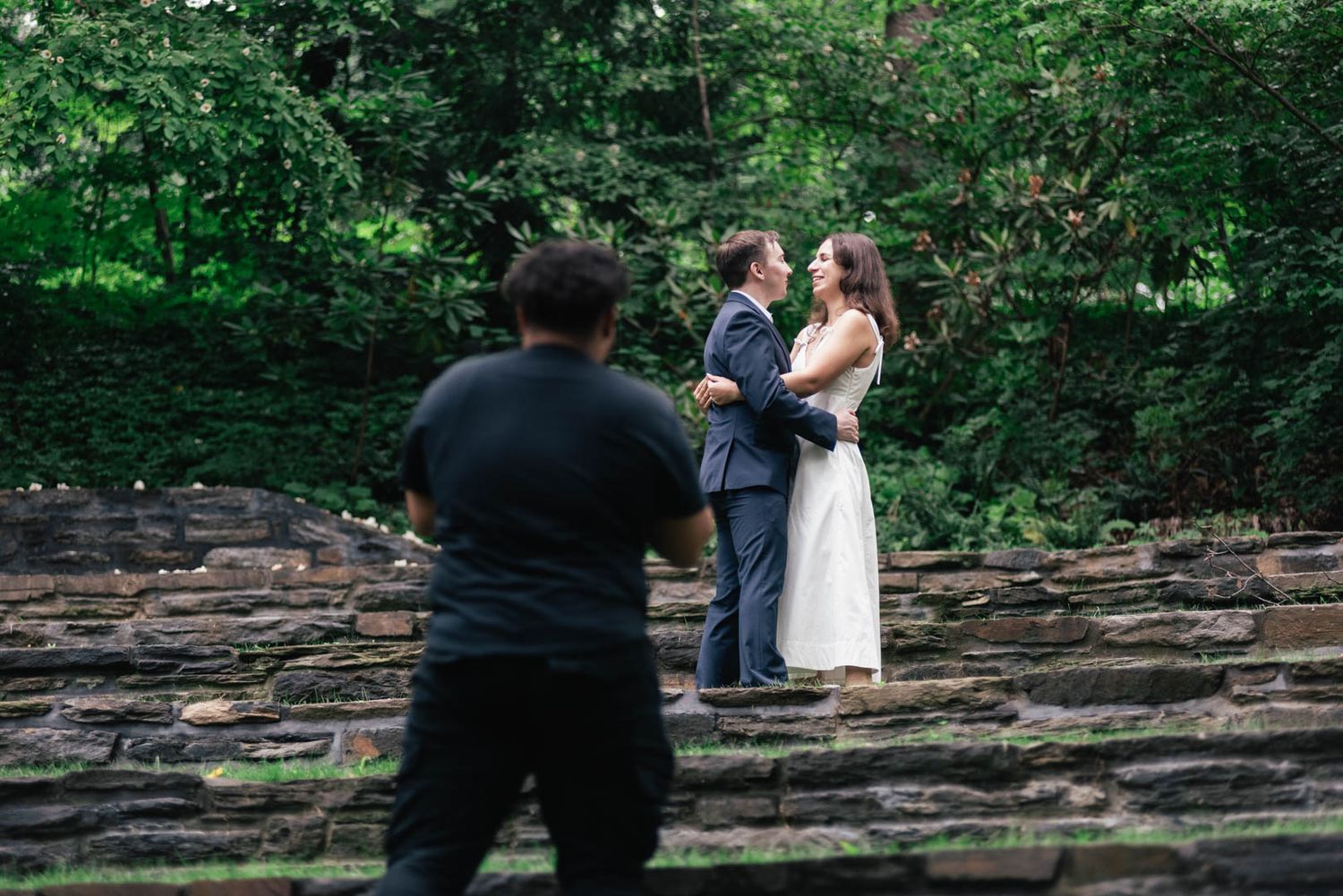 A couple sits together on large stone steps surrounded by lush green forest during a summer photo session.