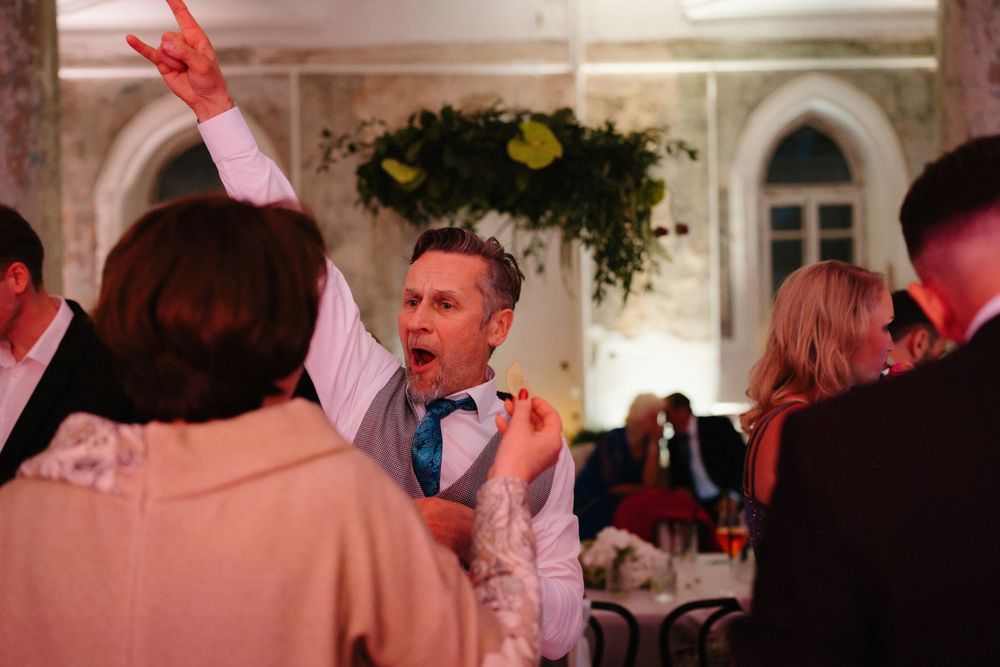 Wedding guests enjoying a lively celebration in an elegant venue with decorative greenery and arched windows.