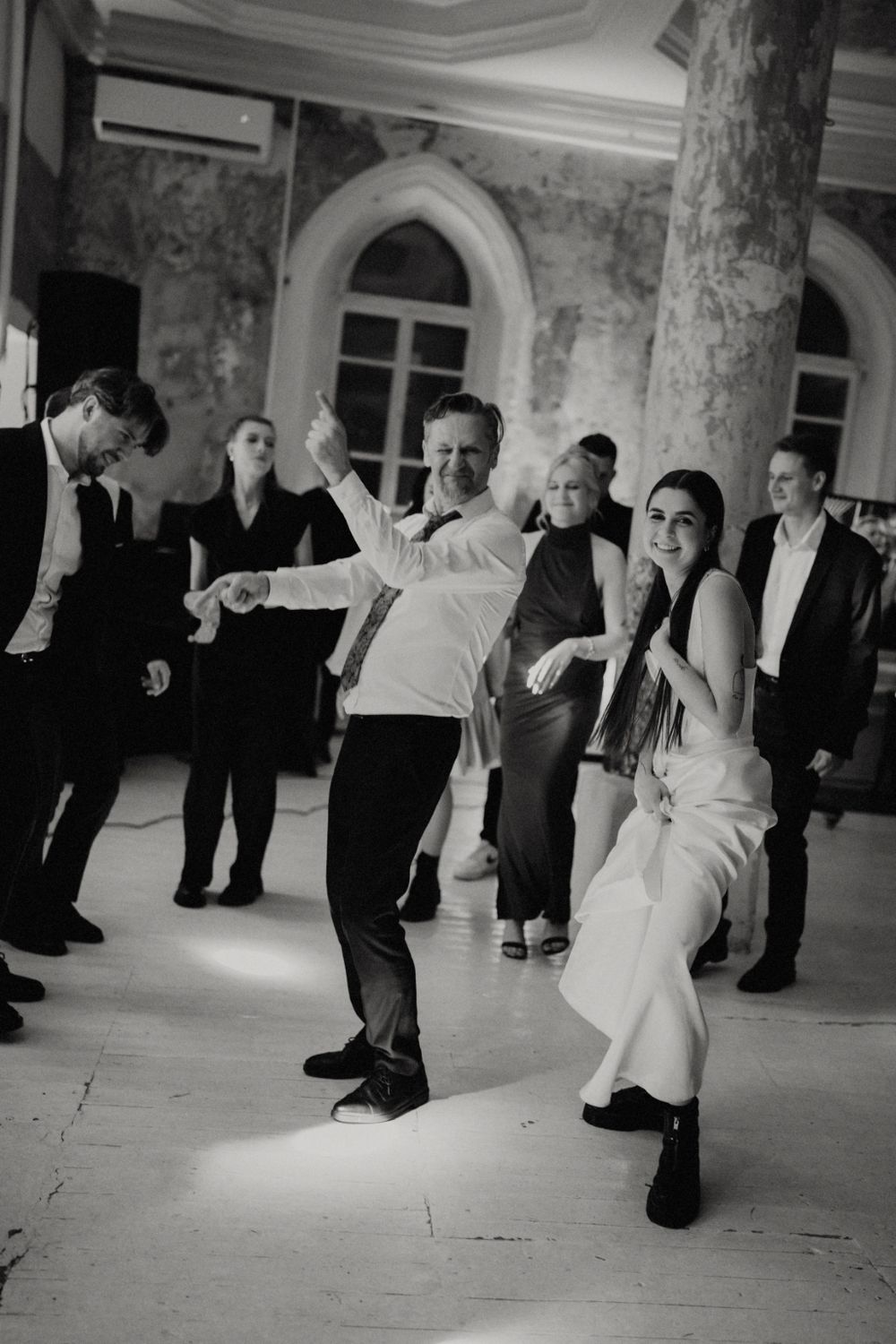 Wedding guests dancing energetically on dance floor in historic venue with arched windows.