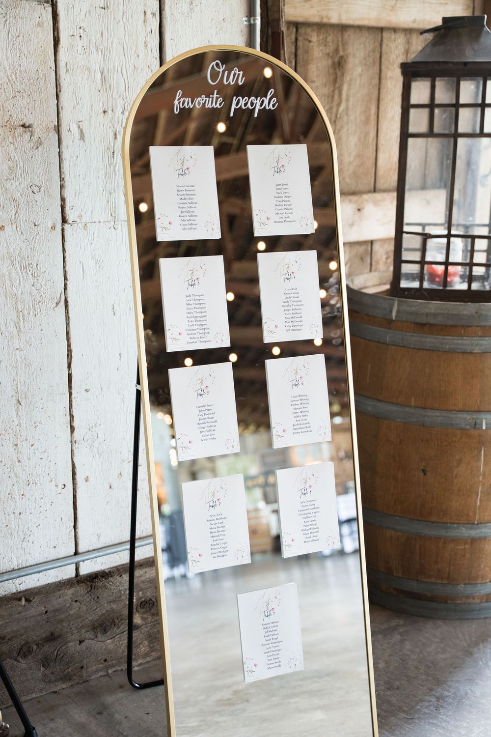 A rustic wedding seating chart displayed on an arched mirror frame next to wooden wine barrels in a barn venue.