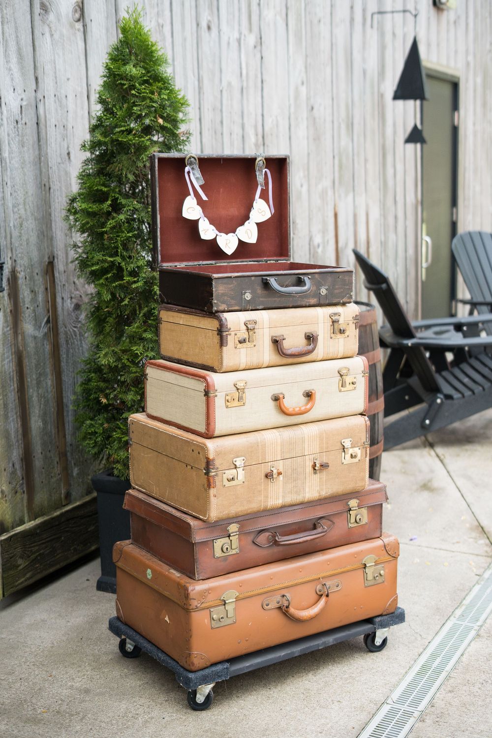 Vintage brown leather suitcases stacked on rolling caster wheels create mobile planter stands against wooden fence.