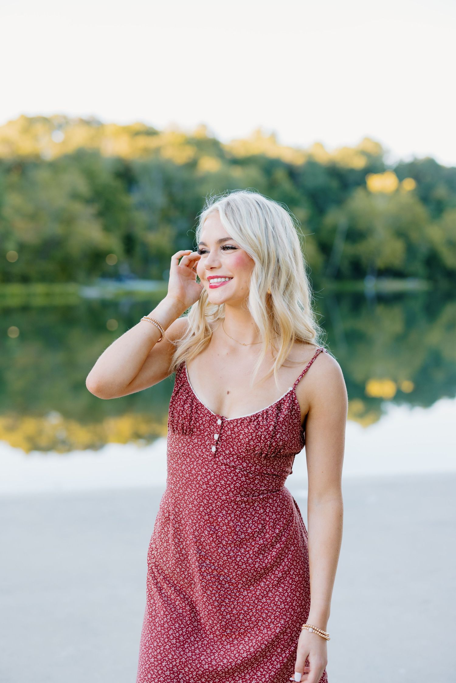 Woman in red floral dress smiling by waterfront during sunset.