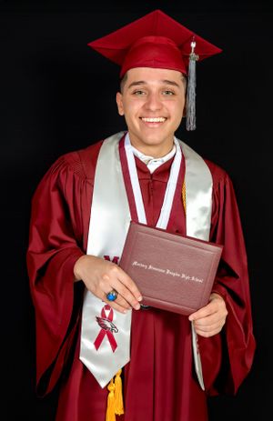 Graduate in red cap and gown holding diploma with honor cords and decorated stole against black background.