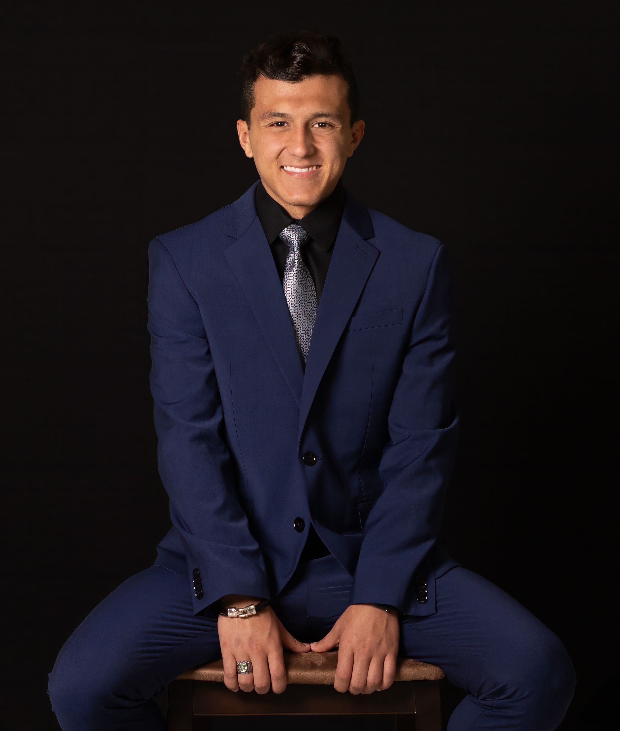 Studio portrait of person in tailored navy suit seated on stool against black background.