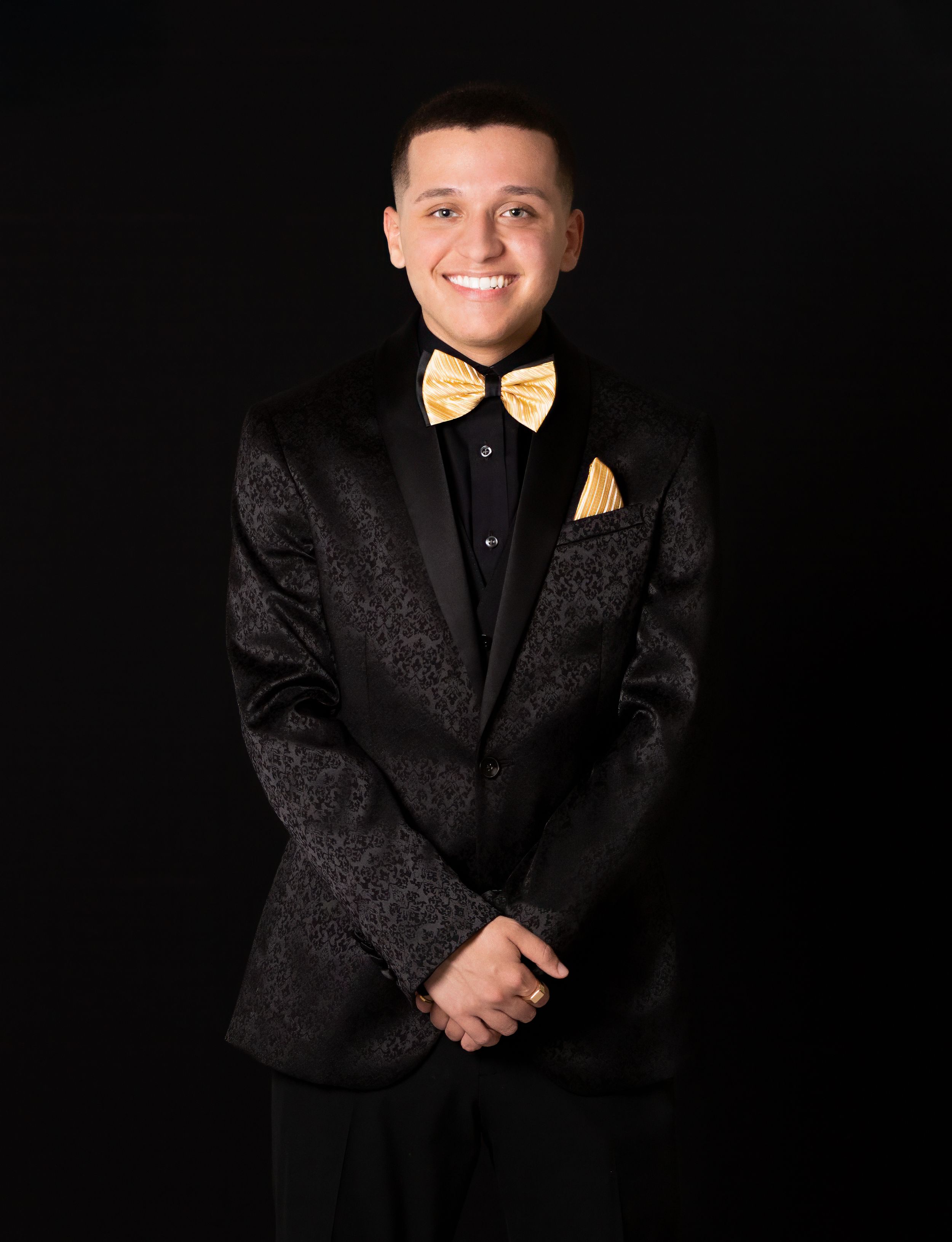 Formal studio portrait showing a person in a black tuxedo with gold bow tie and pocket square against dark background.