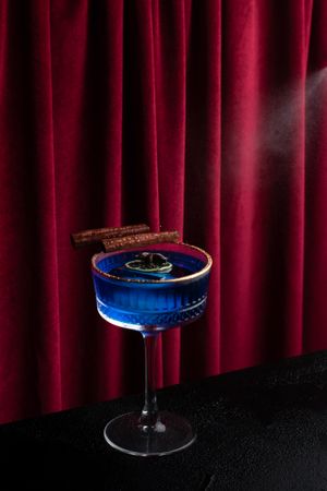 Blue cocktail in martini glass with chocolate garnish against red velvet curtain.