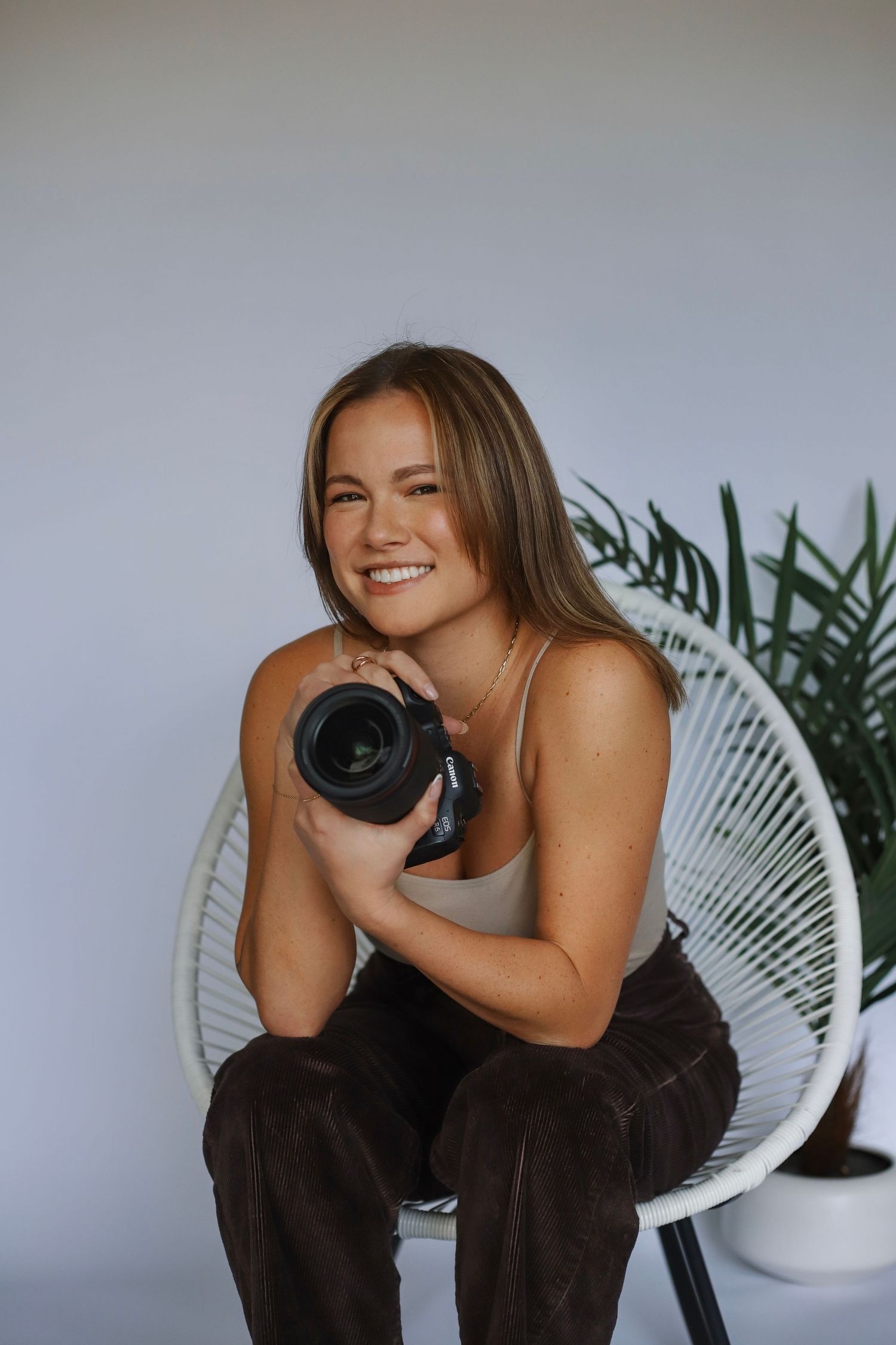 Photographer sitting in white chair with camera and plants in background.