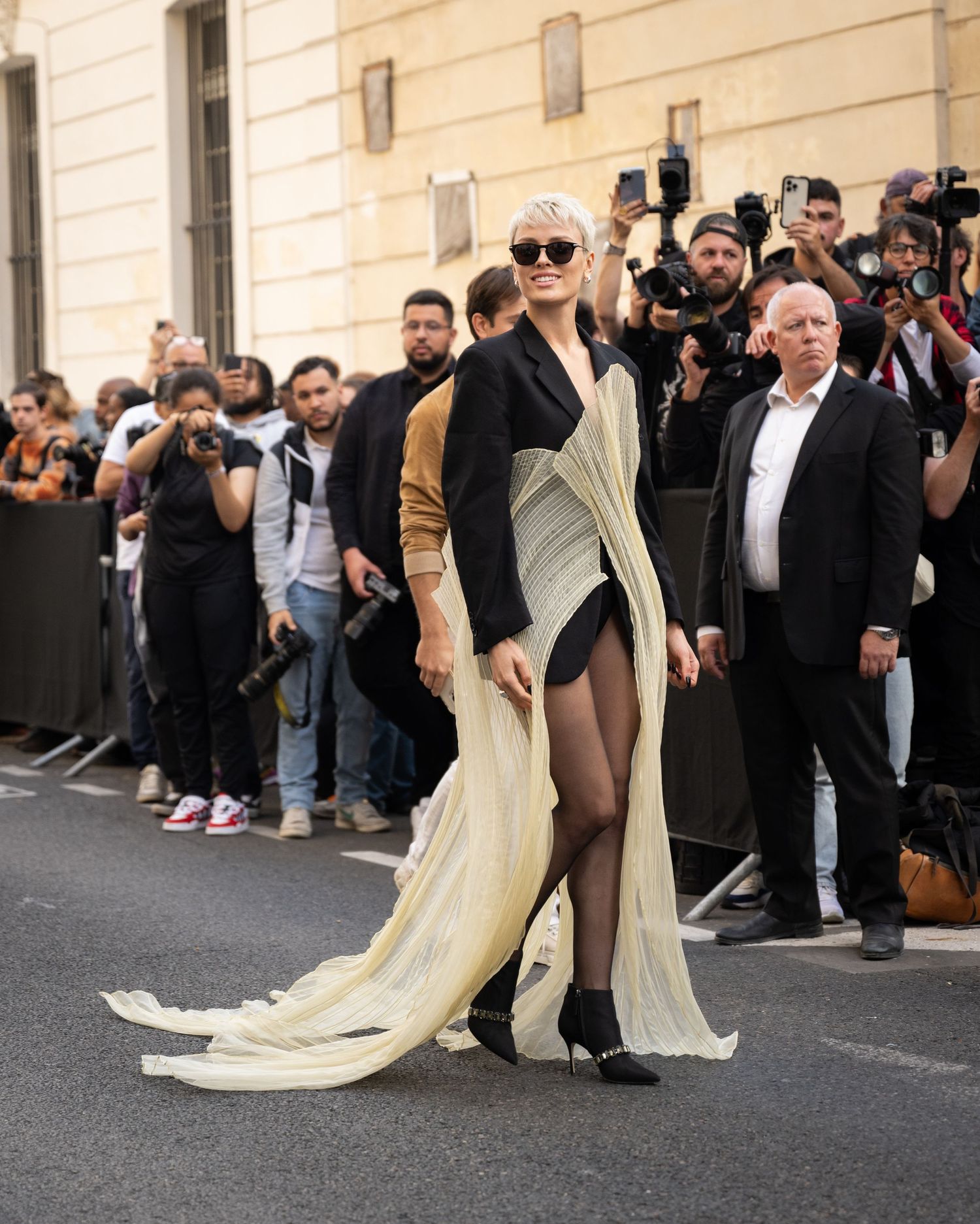 Dramatic cream flowing gown and sunglasses photographed on a street with crowd of onlookers.