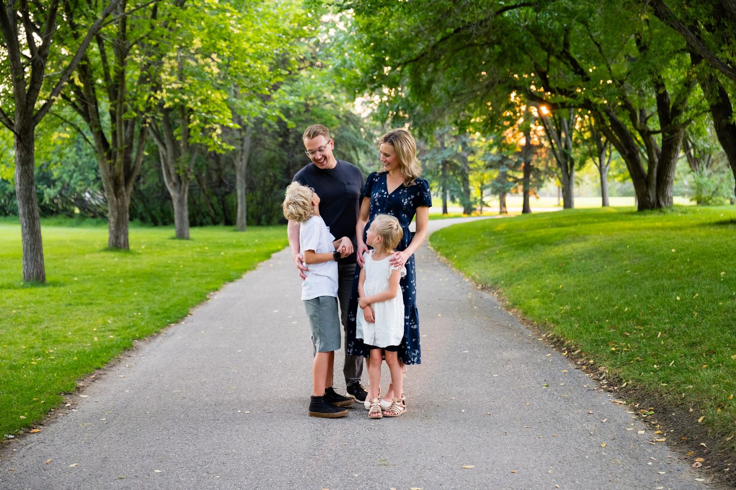 People walk along a tree-lined path in a lush green park on a sunny summer day.
