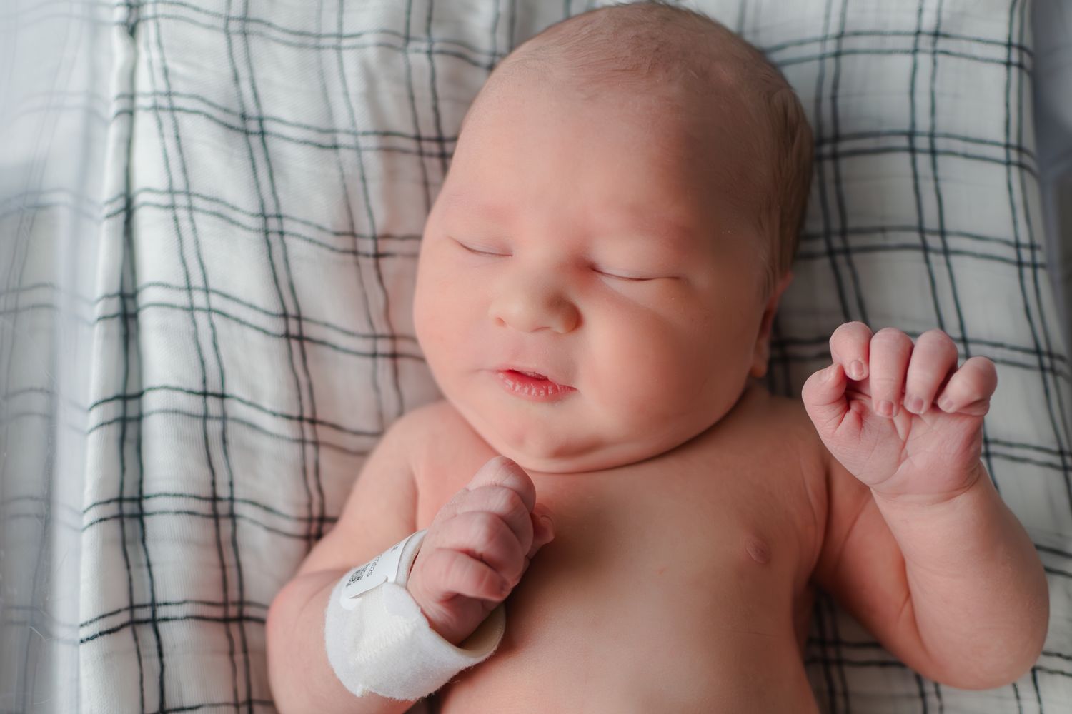 Newborn baby sleeping peacefully on gray plaid blanket with tiny fist raised and bandaged hand.