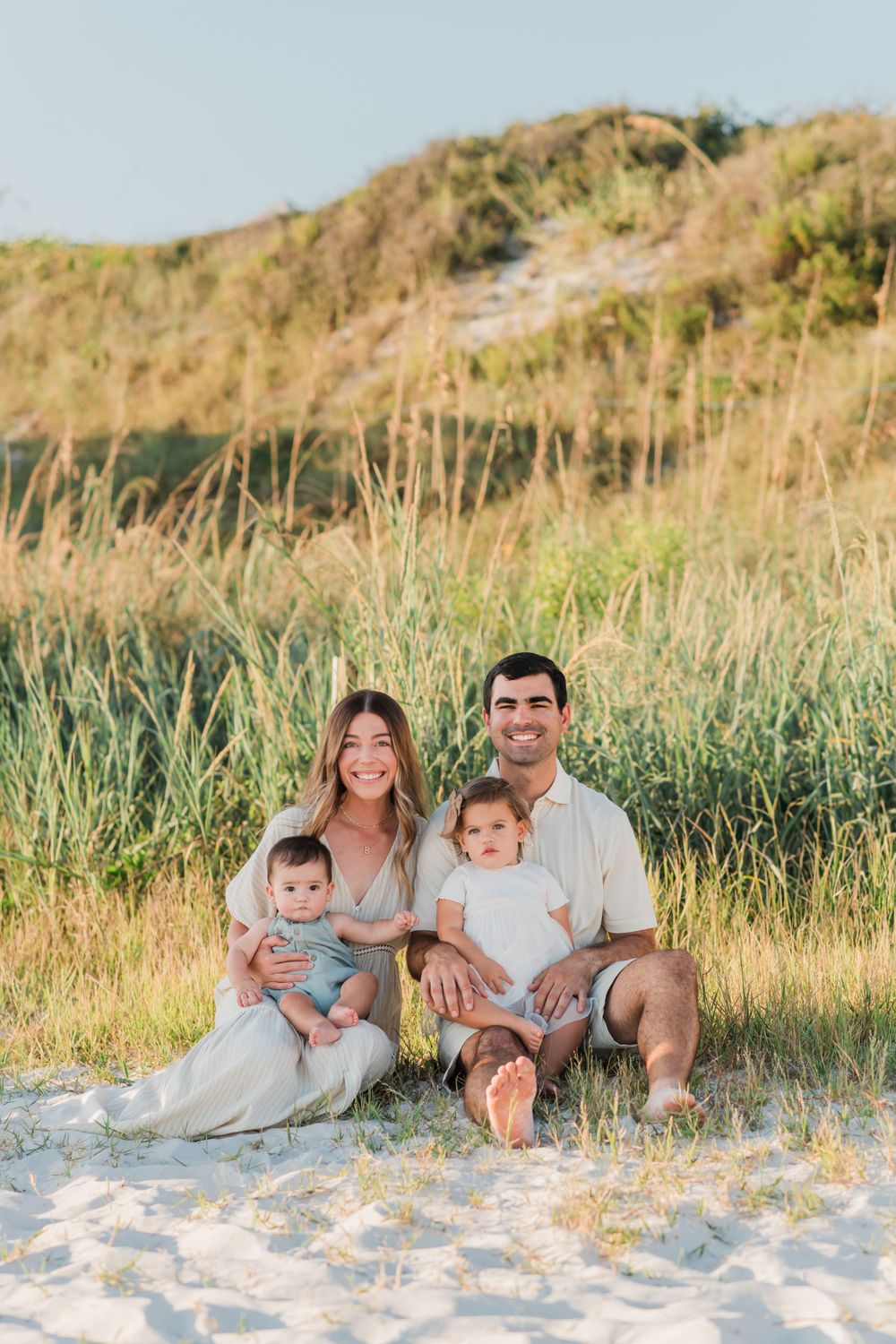 A small family sits together on a sandy beach with tall grass and hills in the background during golden hour.
