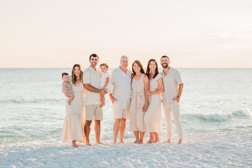 A group in coordinated neutral outfits stands together on a white sandy beach at sunset with ocean waves behind them.