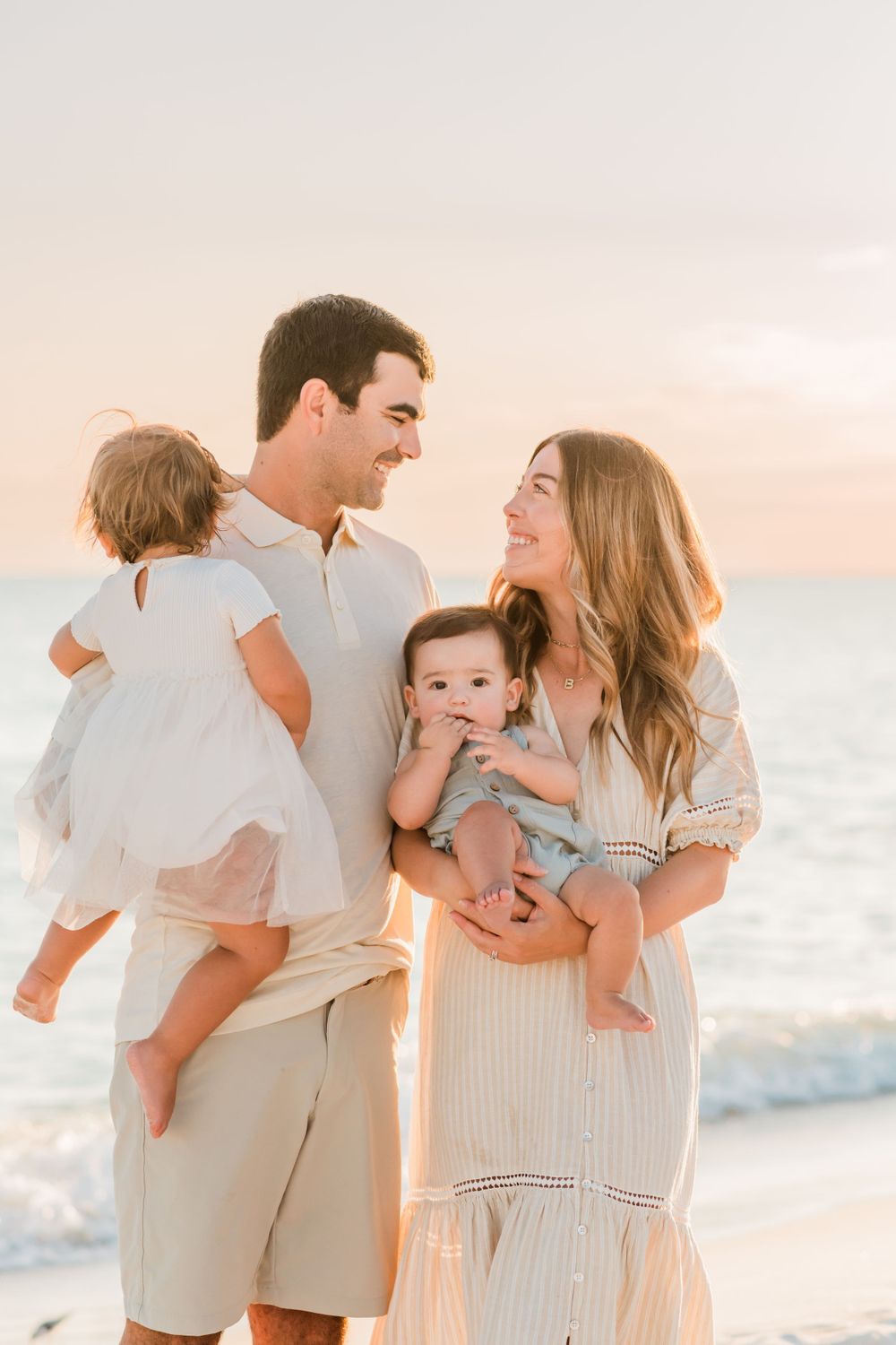 A family wearing light colored clothing smiles at each other while standing on a beach during sunset.