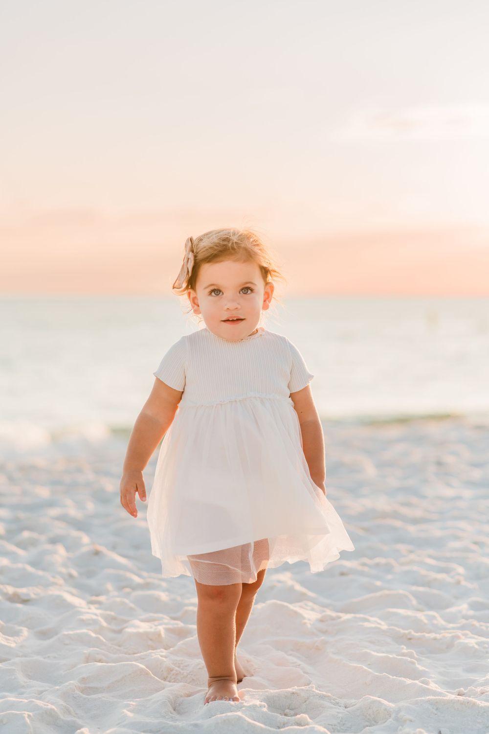 Young child in white dress walking barefoot on sandy beach at sunset with soft pink sky in background.