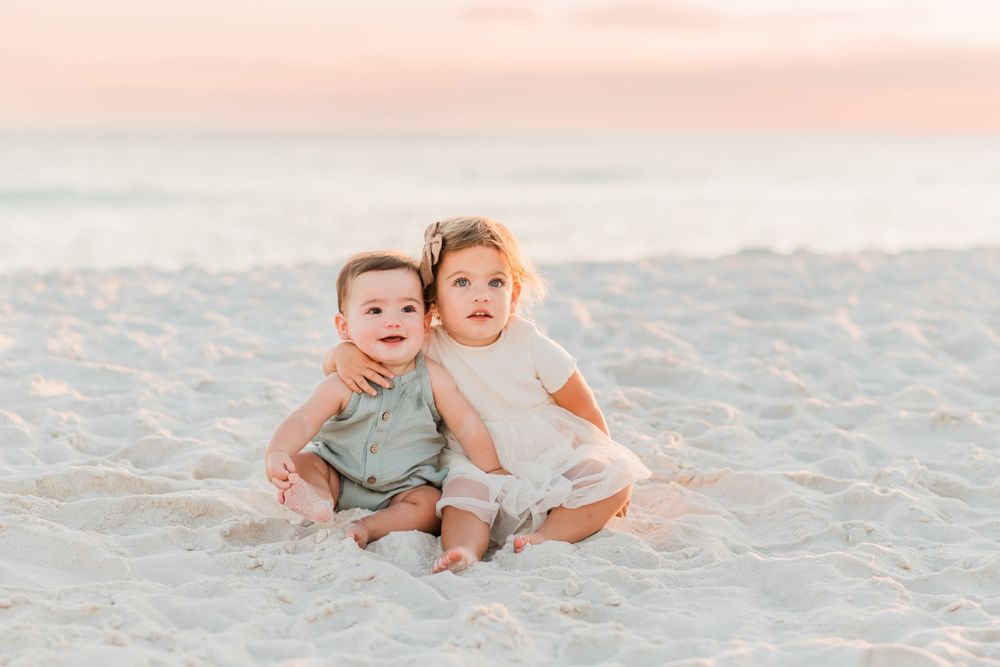 Two young children sit together on white sand beach during sunset with soft pink sky in background.