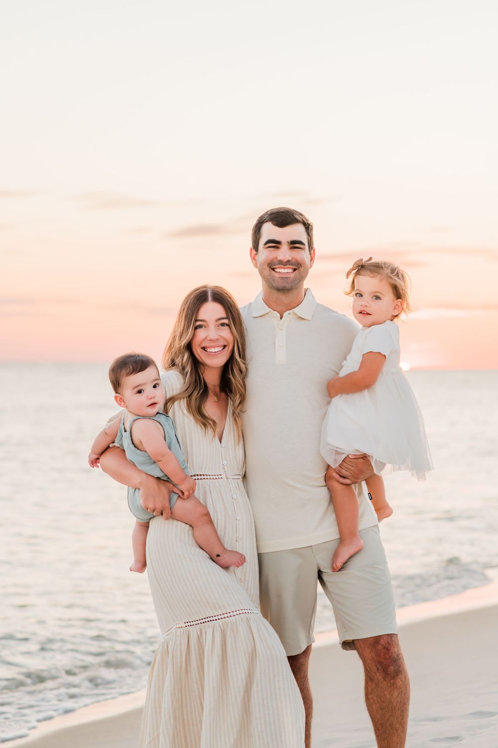 A family of four poses together on a beach at sunset wearing coordinated light colored outfits.
