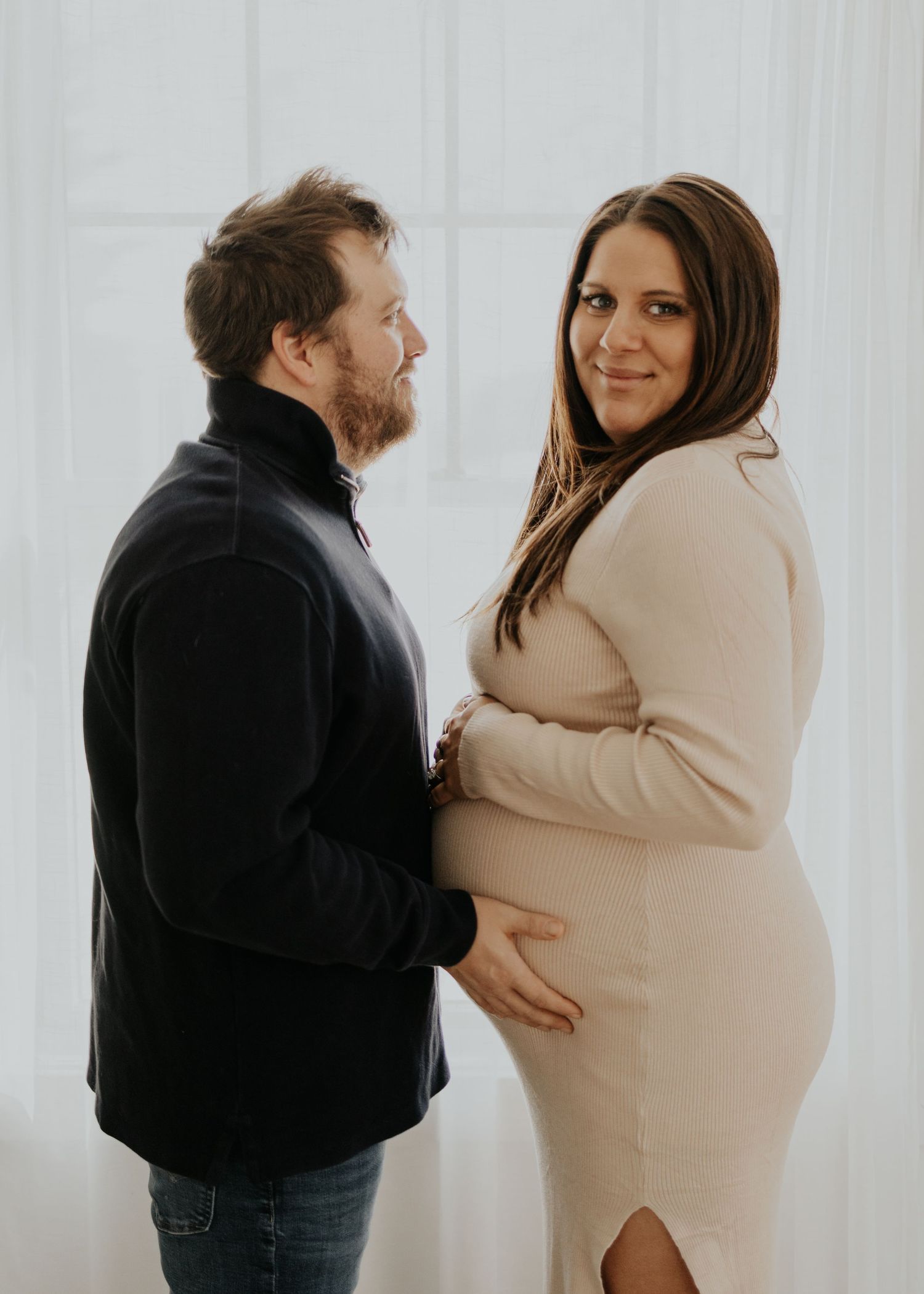 Couple stands close together with person in beige dress showing off pregnant belly while person in black jacket looks on lovingly.