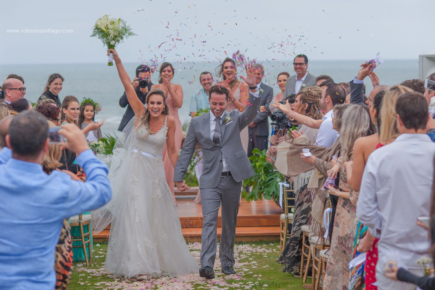 Celebrating couple walk down aisle as guests cheer and throw confetti during outdoor beach wedding ceremony.