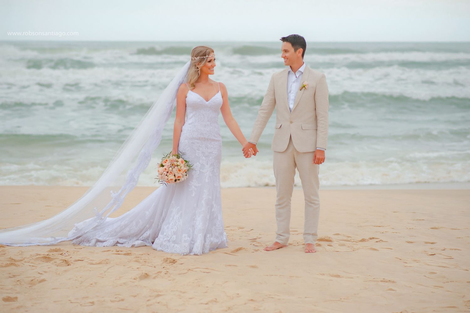 A couple holding hands on a beach during their wedding ceremony with waves crashing in the background.