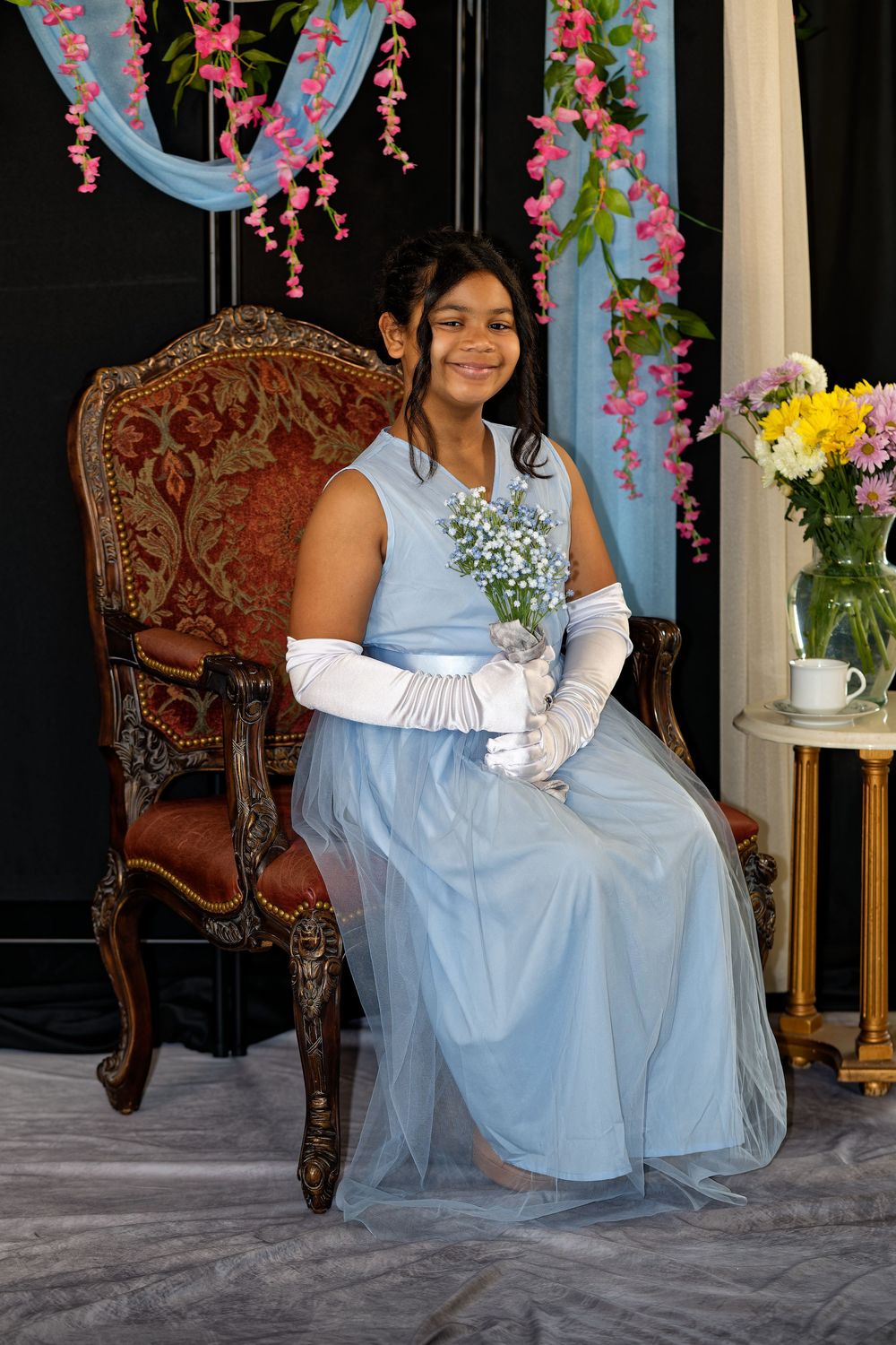 A person in a light blue tulle dress sits elegantly on an ornate antique chair surrounded by pink floral decorations.