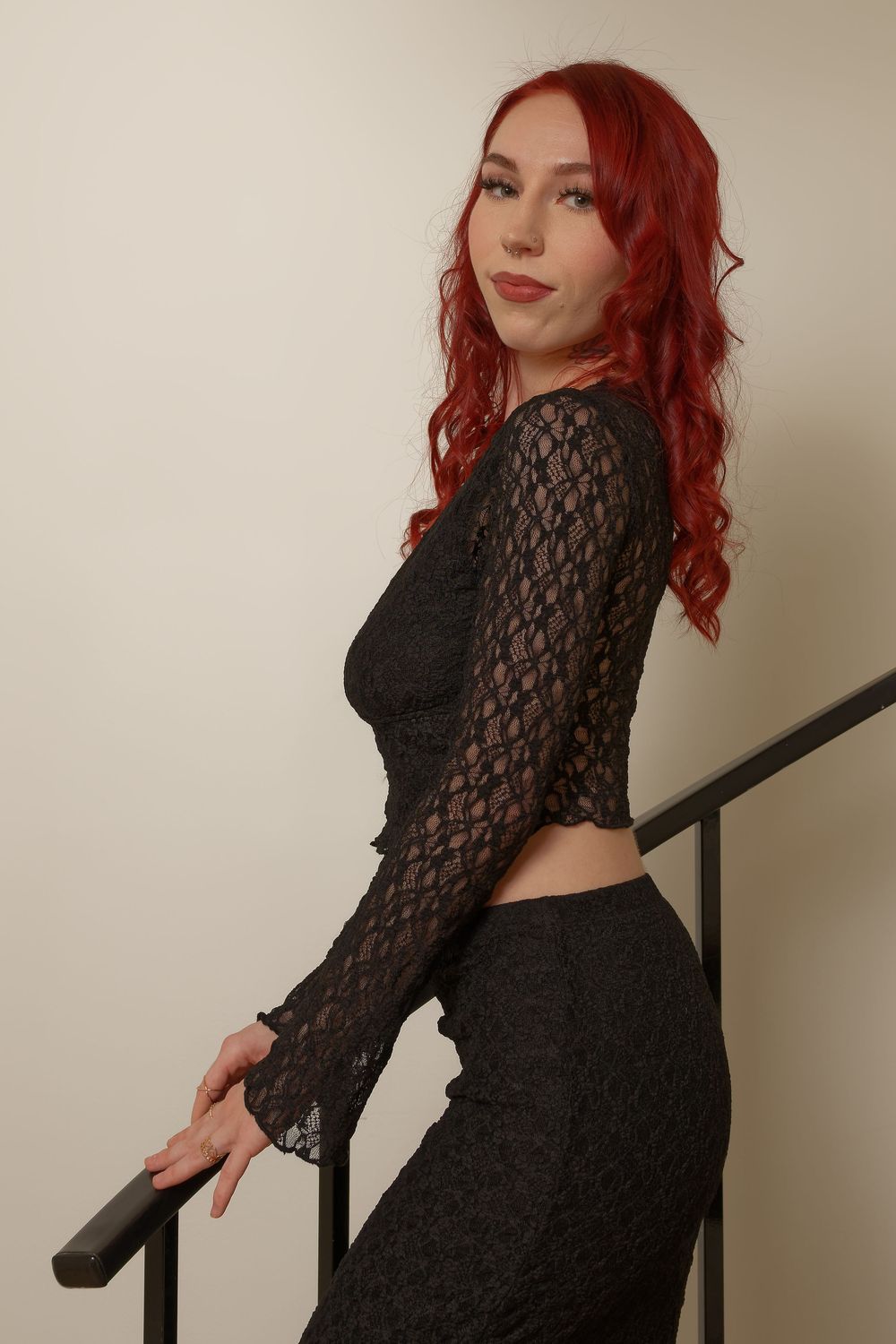 A figure in a black lace dress with vibrant red curly hair poses against a light colored wall in multiple angles.