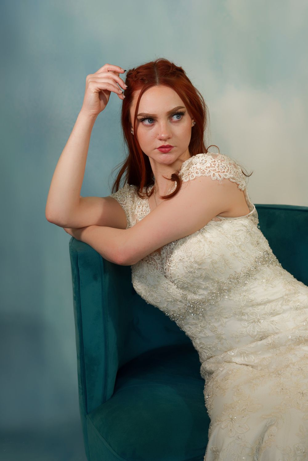A person in a white lace wedding dress poses elegantly on a teal velvet chair in a studio setting.