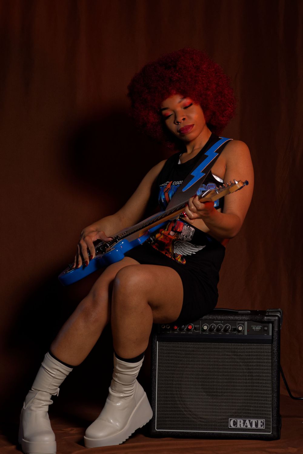 Musician/Model with red afro playing electric guitar while sitting on amplifier.