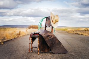 rustic chair sits on rural dirt road with chaps laying across it. teal rope and tan cowboy hat hang off the back.