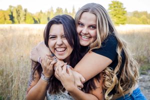 blond girl hugs brunette girl from behind as they smile at the camera surrounded by golden field
