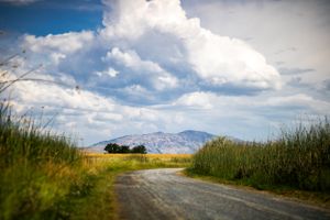 dirt road leads around corner of green fields, mountain can be seen in distance and majestic clouds fill the sky