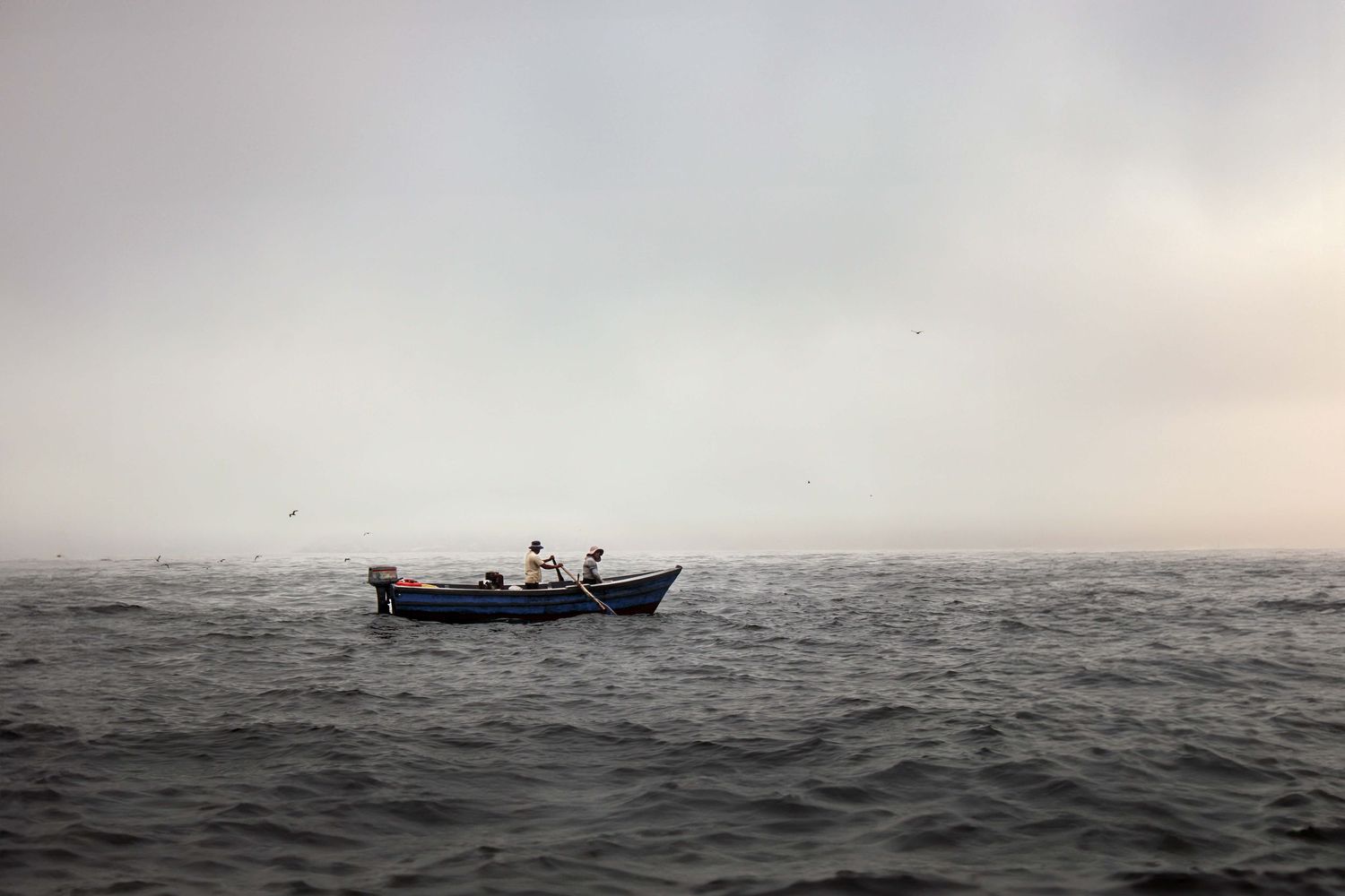 small fishing boat with two men sail through calm ocean waters. it is misty, mysterious, and some birds can be seen far in the background