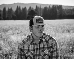 black and white image of serious young man looking at camera out the side of his eye. he is wearing hat, plaid shirt, and in a field